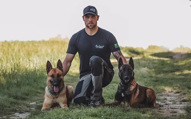Trainer kneeling on a farm path with two shepherd dogs in a sit-stay