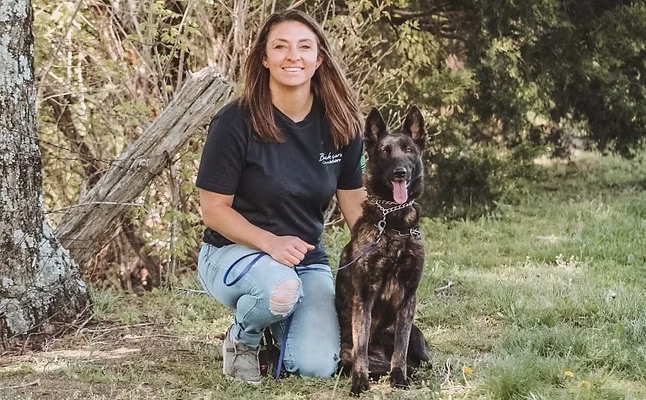 Trainer kneeling beside a dark Belgian Malinois at the treeline