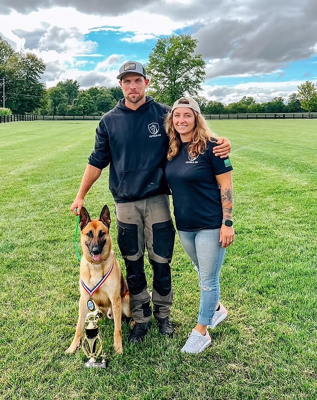 Two trainers on a field with a Belgian Malinois wearing a competition medal