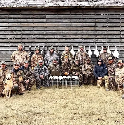 A large group of hunters and a dog in front of a barn with a rack of snow geese