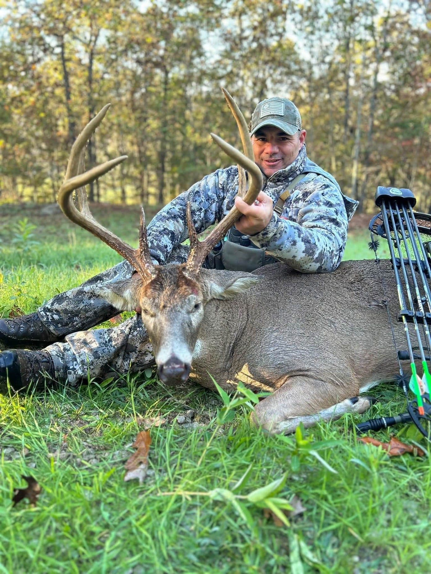 The author kneeling in the grass behind a large whitetail buck, holding its antlers, with a bow propped beside it.