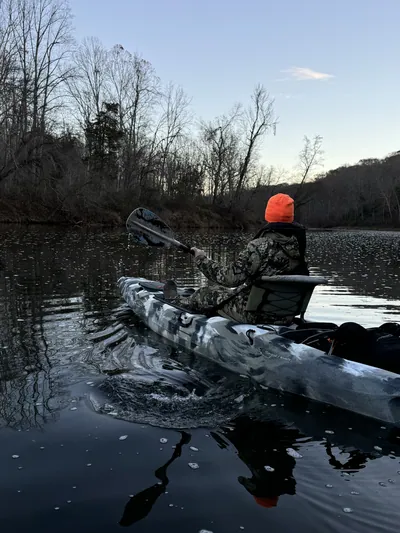 A kayaker in camouflage paddles across a still river at dusk.