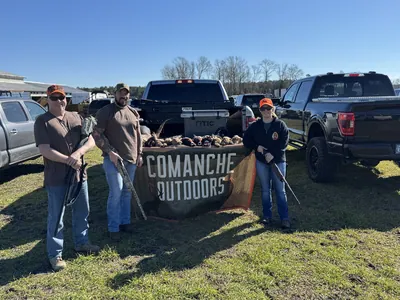 Three hunters stand by a Comanche Outdoors banner at a pheasant hunt tailgate.