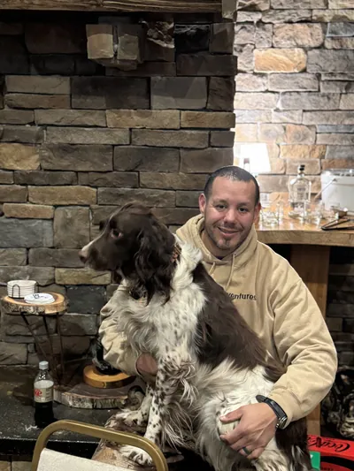 A man smiles in a lodge with a springer spaniel on his lap.