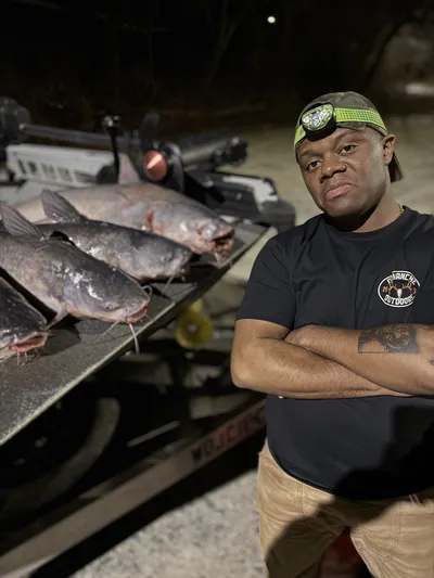 An angler holds a catfish at night by the light of a headlamp.
