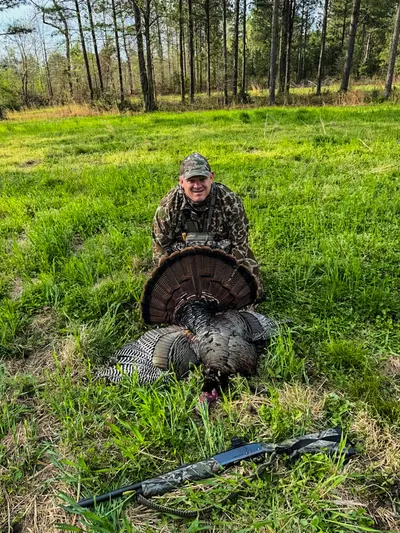 A smiling hunter kneels behind a turkey with its tail fanned.