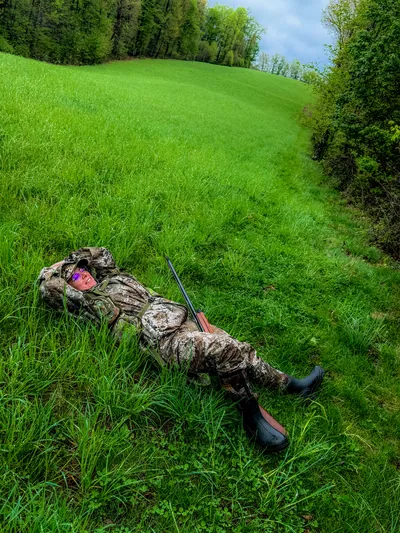 A turkey hunter in a face mask sits on a green slope.