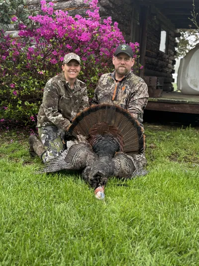 A couple kneels with a wild turkey in front of pink azaleas.