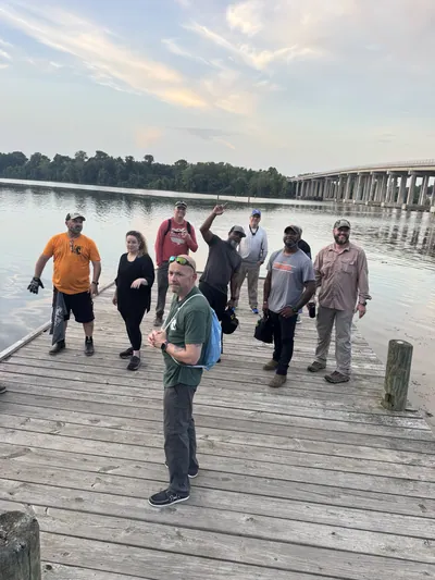 A group of participants stands on a wooden dock by a river at dusk.