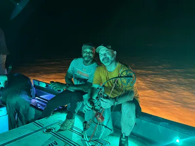 Two men sit on a bowfishing boat lit by teal and orange lights at night.