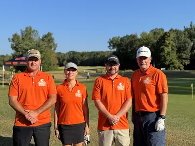 Four golfers in orange Comanche Outdoors polos stand on a course under a blue sky.