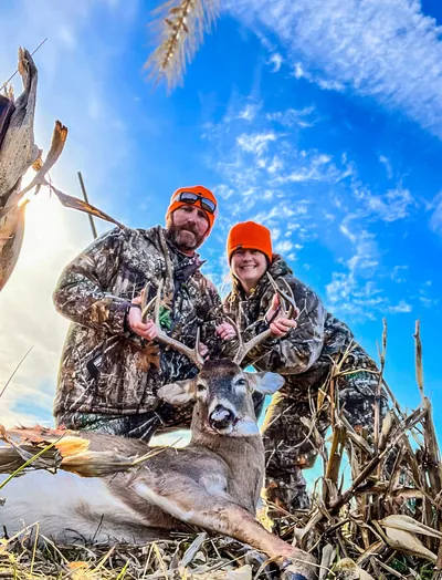 A couple poses with a whitetail buck under a dramatic sky.