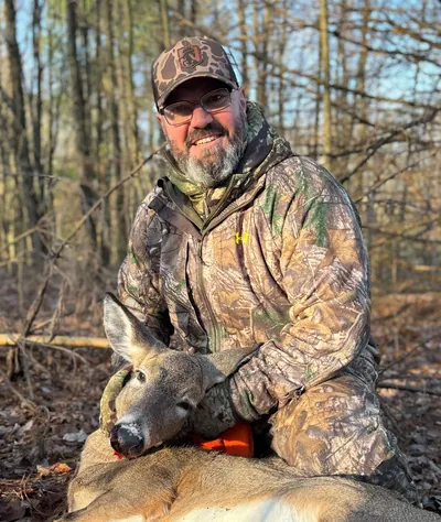 A hunter in a Comanche Outdoors cap poses with a doe in warm evening light.