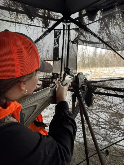A hunter aims from a box blind, seen over his shoulder.