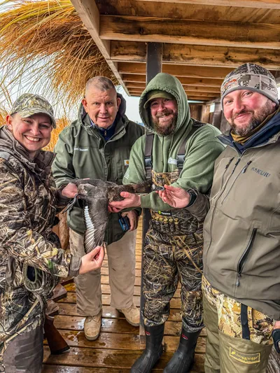 Four hunters in camouflage smile in a duck blind, one holding a duck.