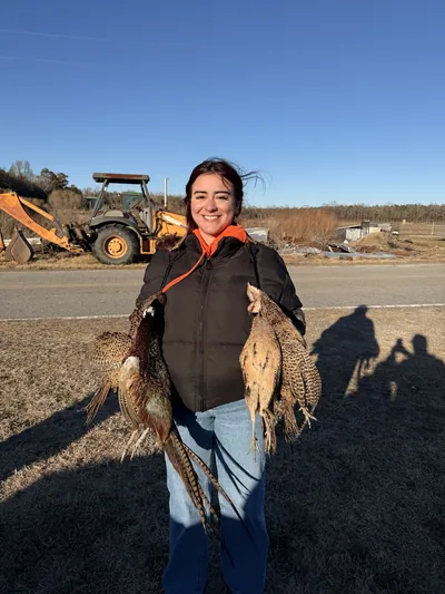 A smiling hunter holds pheasants, with a backhoe behind.
