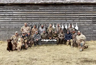 About two dozen hunters and two Labradors pose in front of a weathered barn.