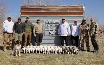 A hunting group with a springer spaniel and the day's geese beside an outfitter's sign.