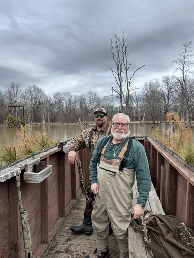 Two hunters in waders stand in a wooden duck blind over flooded timber.