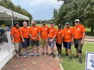 Volunteers in orange Comanche Outdoors polos stand together at a golf course.