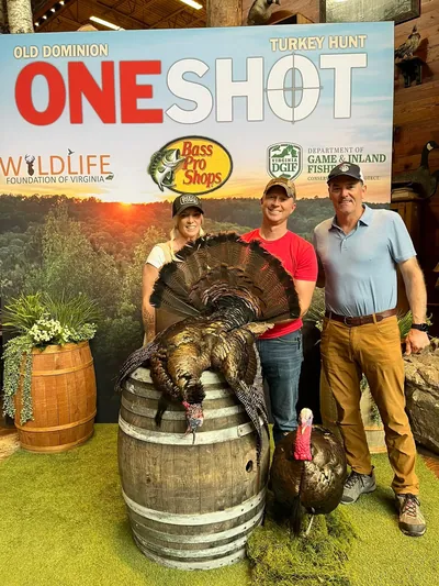 Three people pose in front of a sponsor backdrop at a turkey hunting event.