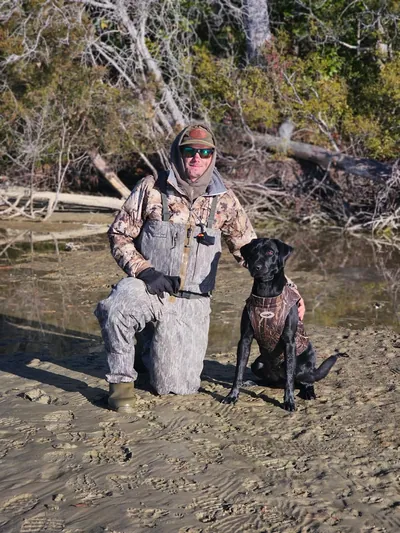 A handler and a black Labrador on a sandy shore.